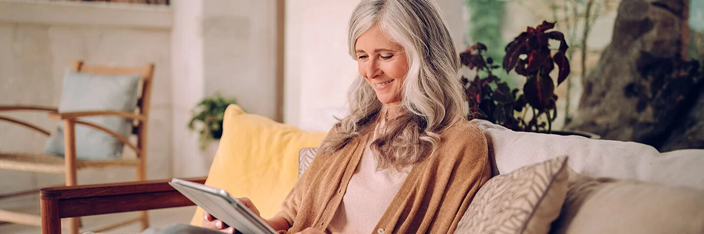 Eine ältere Frau mit langen grauen Haaren sitzt entspannt auf einem gemütlichen Sofa in einem hellen Wohnzimmer. Sie trägt einen beigefarbenen Cardigan und hält ein Tablet in den Händen, auf das sie lächelnd schaut. Im Hintergrund sind Pflanzen, Regale und ein Sessel zu sehen, was dem Raum eine einladende Atmosphäre verleiht.