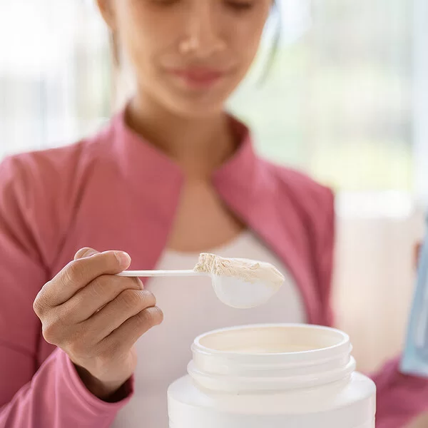 Eine junge Frau entnimmt mit einem Dosierlöffel eine Portion weißes Pulver aus einer Plastikdose. In der anderen Hand hält sie eine Trinkflasche mit Wasser.