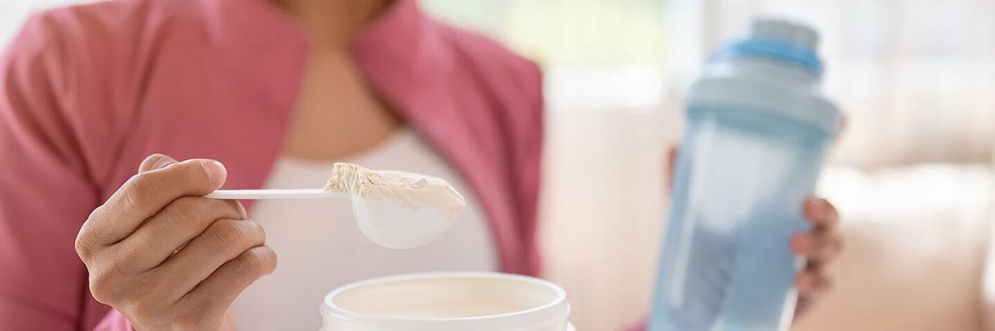 Eine junge Frau entnimmt mit einem Dosierlöffel eine Portion weißes Pulver aus einer Plastikdose. In der anderen Hand hält sie eine Trinkflasche mit Wasser.