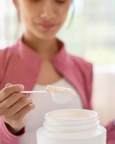 Eine junge Frau entnimmt mit einem Dosierlöffel eine Portion weißes Pulver aus einer Plastikdose. In der anderen Hand hält sie eine Trinkflasche mit Wasser.