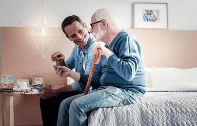 Ein Mann im Jeanshemd hält einen Geschenkumschlag und spricht freundlich seinem alten Vater zugewandt. Der Vater hat trägt eine große Brille, einen blauen Pullover und hält einen Gehstock. Sie sitzen auf der Bettkante im Zimmer des Vaters. 