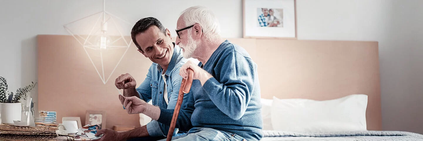 Ein Mann im Jeanshemd hält einen Geschenkumschlag und spricht freundlich seinem alten Vater zugewandt. Der Vater hat trägt eine große Brille, einen blauen Pullover und hält einen Gehstock. Sie sitzen auf der Bettkante im Zimmer des Vaters. 