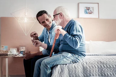 Ein Mann im Jeanshemd hält einen Geschenkumschlag und spricht freundlich seinem alten Vater zugewandt. Der Vater hat trägt eine große Brille, einen blauen Pullover und hält einen Gehstock. Sie sitzen auf der Bettkante im Zimmer des Vaters. 