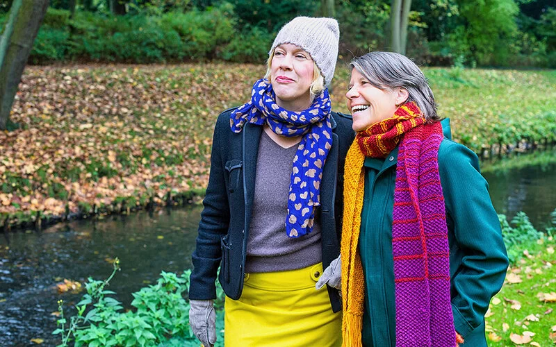 Zwei Frauen in herbstlicher Kleidung gehen, umgeben von Bäumen, in der Nähe eines Gewässers spazieren.