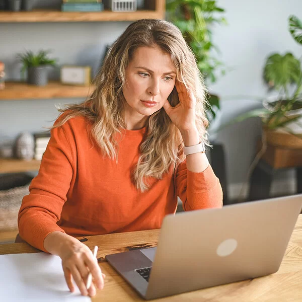 Eine Frau sitzt mit müdem Blick vor einem Laptop, den Kopf mit einer Hand gestützt.