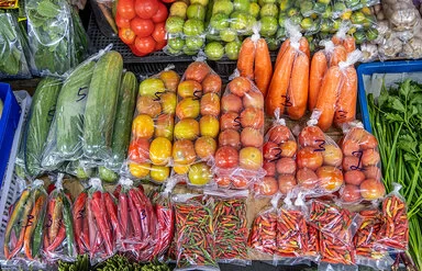 Nahaufnahme der Auslage eines Gemüsemarktstandes mit unter anderem Gurken, Tomaten, Karotten und Peperoni. Das Gemüse wird in Kunststoffverpackungen angeboten.