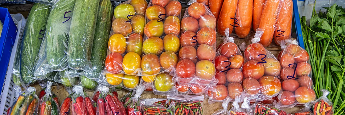 Nahaufnahme der Auslage eines Gemüsemarktstandes mit unter anderem Gurken, Tomaten, Karotten und Peperoni. Das Gemüse wird in Kunststoffverpackungen angeboten.