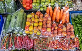 Nahaufnahme der Auslage eines Gemüsemarktstandes mit unter anderem Gurken, Tomaten, Karotten und Peperoni. Das Gemüse wird in Kunststoffverpackungen angeboten.