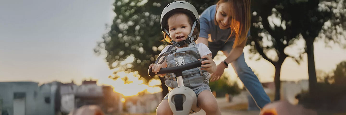 Ein Kind mit einem Helm auf dem Kopf sitzt auf einem kleinen Fahrrad und wird von der Mutter angeschoben.