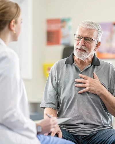 Eine älterer Mann mit Brille und Vollbart spricht beim Check-up mit seiner Ärztin über seine Herzbeschwerden.