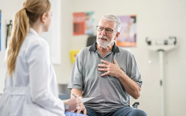 Eine älterer Mann mit Brille und Vollbart spricht beim Check-up mit seiner Ärztin über seine Herzbeschwerden.