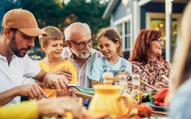 Ein älteres Paar, zwei Kinder, ein junger Mann und weitere Personen sitzen fröhlich feiernd an einem Esstisch im Garten.