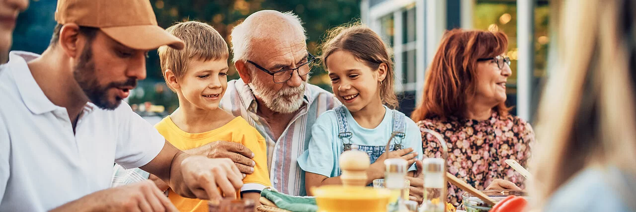 Ein älteres Paar, zwei Kinder, ein junger Mann und weitere Personen sitzen fröhlich feiernd an einem Esstisch im Garten.