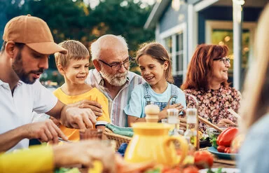 Ein älteres Paar, zwei Kinder, ein junger Mann und weitere Personen sitzen fröhlich feiernd an einem Esstisch im Garten.