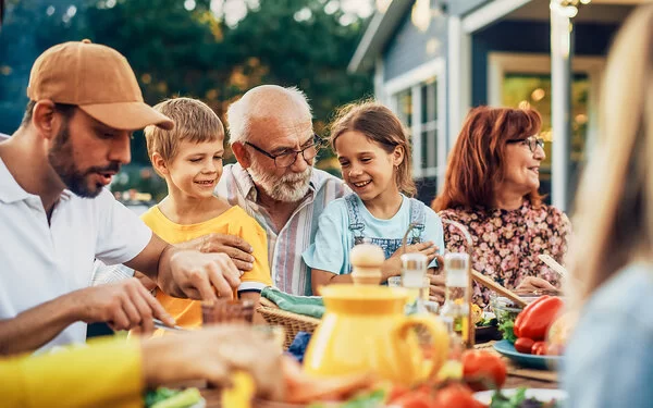 Ein älteres Paar, zwei Kinder, ein junger Mann und weitere Personen sitzen fröhlich feiernd an einem Esstisch im Garten.