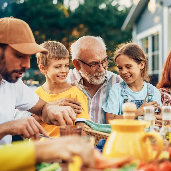 Ein älteres Paar, zwei Kinder, ein junger Mann und weitere Personen sitzen fröhlich feiernd an einem Esstisch im Garten.