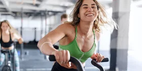 Eine junge Frau in grünem Sport-Oberteil trainiert lächelnd auf einem Fahrrad-Ergometer im Fitnessstudio.