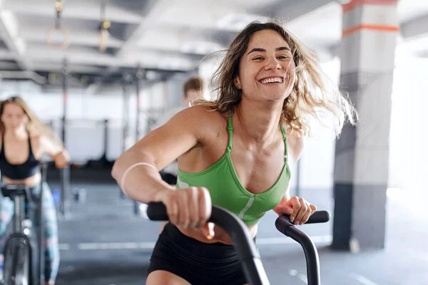 Eine junge Frau in grünem Sport-Oberteil trainiert lächelnd auf einem Fahrrad-Ergometer im Fitnessstudio.