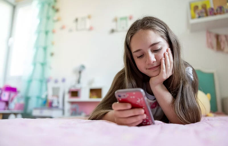 Ein junges Mädchen liegt auf einem Bett und schaut lächelnd auf ihr Smartphone.