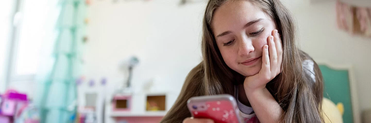 Ein junges Mädchen liegt auf einem Bett und schaut lächelnd auf ihr Smartphone.