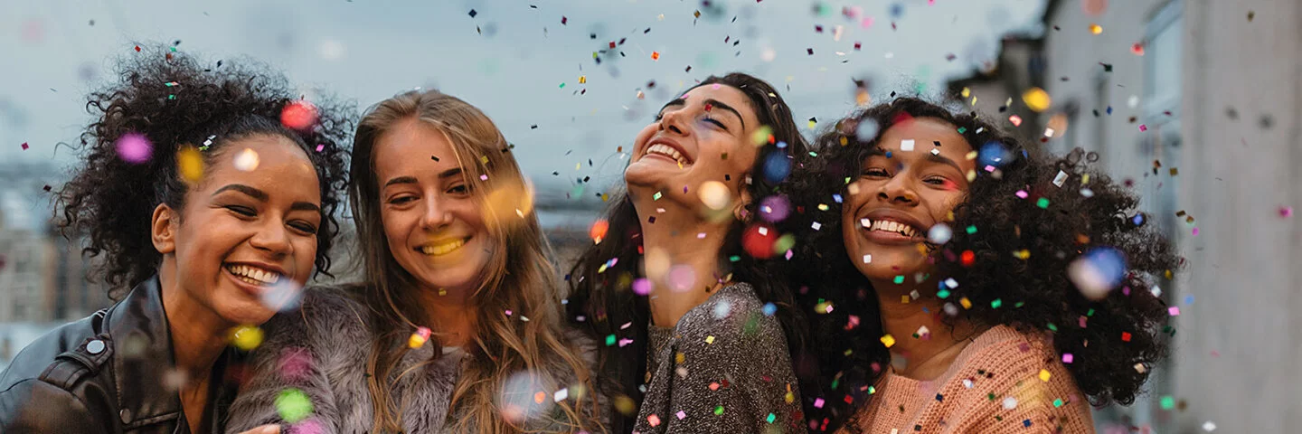 Vier lachende junge Frauen stehen im Dämmerlicht auf einer Terrasse im Konfettiregen.