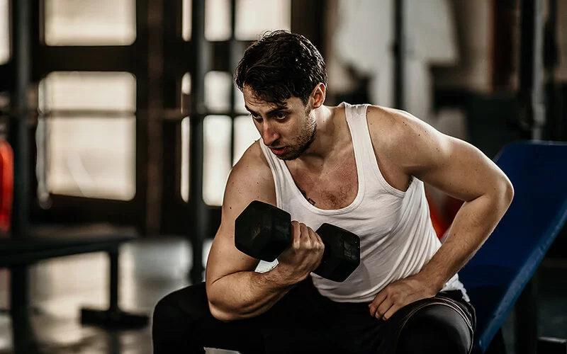 Ein Mann im ärmellosen Sportshirt sitzt im Fitnessstudio auf einer Bank und trainiert mit einer Kurzhantel. Er macht Armbeugen.