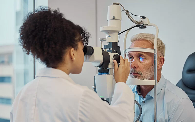 Eine Augenärztin oder eine medizinische Fachangestellte untersucht das linke Auge eines älteren Mannes mit einem augenärztlichen Messgerät.