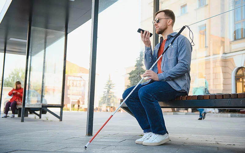 Ein sehbehinderter junger Mann mit dunkler Sonnenbrille sitzt in der Stadt auf einer Bank. In der linken Hand hält er einen weißen Langstock, in der rechten ein Smartphone auf der Höhe seines Kinns.