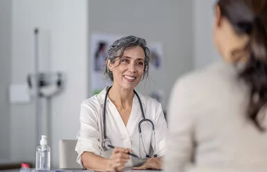 Eine Ärztin spricht mit einer dunkelhaarigen Patientin. Sie trägt eine weiße Bluse und ein Stethoskop um den Hals und lächelt die Patientin freundlich an. Im Hintergrund ist ein Raum in einer Arztpraxis zu sehen.