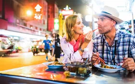 Eine Frau und ein Mann, um die 40 Jahre alt, sitzen nebeneinander in einem chinesischen Street-Food-Restaurant und essen mit Stäbchen. Die Frau gibt dem Mann etwas zum Probieren in den Mund und lacht dabei.