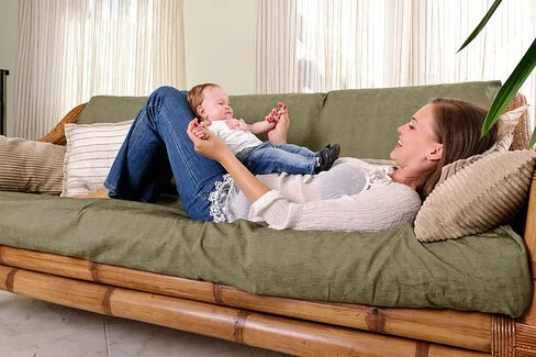 Eine Mutter liegt mit ihrem Baby auf einem Sofa mit grüner Polsterung und Bambusgestell.