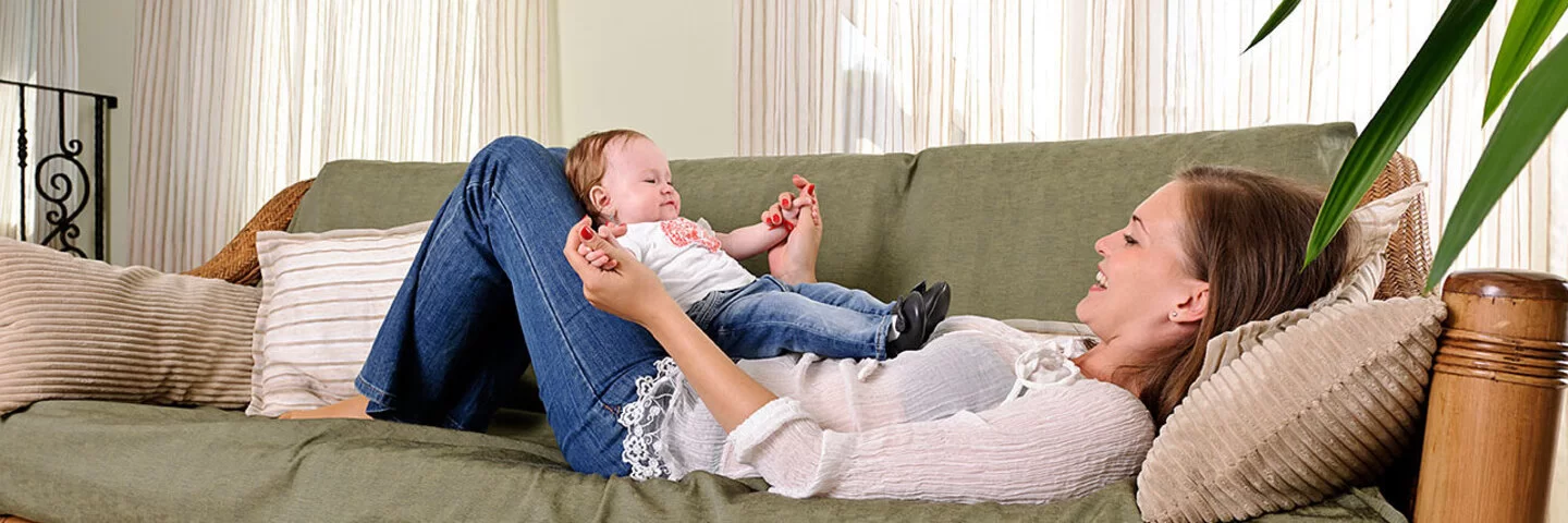 Eine Mutter liegt mit ihrem Baby auf einem Sofa mit grüner Polsterung und Bambusgestell.