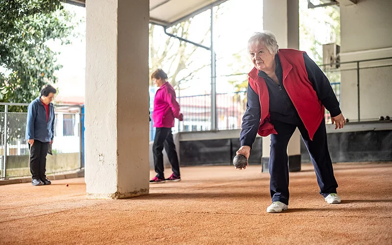 Eine ältere Frau mit roter Weste und silbergrauen Haaren beugt sich nach vorne. In der rechten Hand hält sie eine dunkelgraue Pétanque-Kugel, um sie aus dem Stand zu werfen. Das Spielfeld ist draußen unter der Überdachung neben einem Wohngebäude. Im Hintergrund sind zwei Frauen zu sehen. Sie schauen sich eine kleine Kugel an, die auf dem Spielfeld liegt.