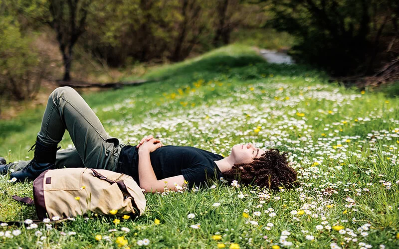 Ein junger Mann mit dunklen Locken, schwarzem T-Shirt und schwarzen Jeans liegt entspannt mit geschlossenen Augen rücklings im Gras. Um ihn herum blühen Gänseblümchen und Löwenzahn.