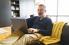 Ein Mann Mitte 50 sitzt entspannt mit einem Laptop auf dem Schoß auf einem Sofa.