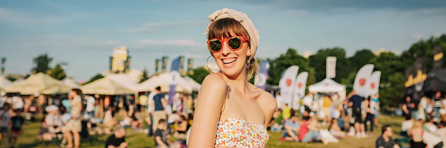 Eine junge Frau steht mit bauchfreiem Top, Sonnenbrille und um den Kopf gewickeltem Tuch auf der Wiese eines sommerlichen Open Airs. Unscharf sind im Hintergrund andere Besucher und mit Zelten überdachte Verkaufsstände zu erkennen.