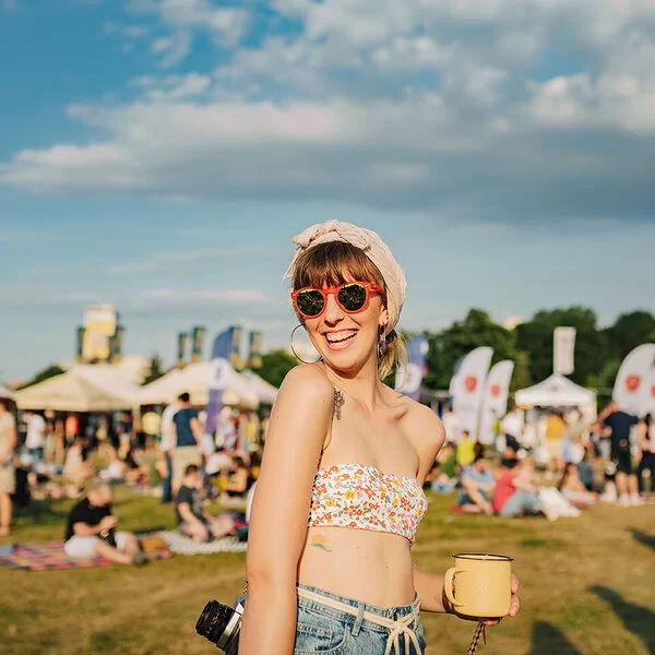 Eine junge Frau steht mit bauchfreiem Top, Sonnenbrille und um den Kopf gewickeltem Tuch auf der Wiese eines sommerlichen Open Airs. Unscharf sind im Hintergrund andere Besucher und mit Zelten überdachte Verkaufsstände zu erkennen.