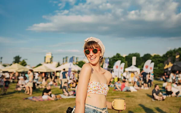 Eine junge Frau steht mit bauchfreiem Top, Sonnenbrille und um den Kopf gewickeltem Tuch auf der Wiese eines sommerlichen Open Airs. Unscharf sind im Hintergrund andere Besucher und mit Zelten überdachte Verkaufsstände zu erkennen.