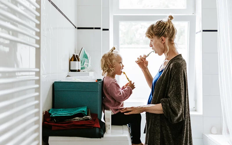 Eine Mutter putzt mit ihrer Tochter im Kindergartenalter im Badezimmer die Zähne. Das Kind sitzt auf der Waschmaschine, die Mutter steht ihm gegenüber. Sie schauen sich in die Augen.