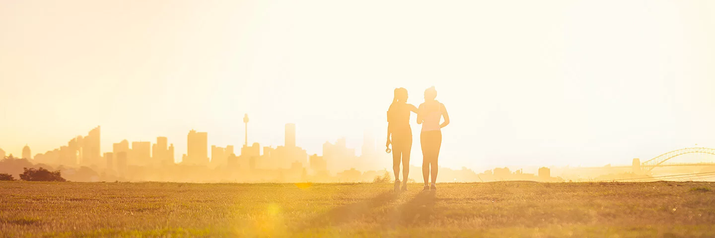Silhouette zweier Frauen, die bei Sonnenuntergang in einem Park spazieren. Im Hintergrund ist die Skyline einer Stadt zu erkennen.