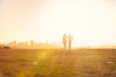 Silhouette zweier Frauen, die bei Sonnenuntergang in einem Park spazieren. Im Hintergrund ist die Skyline einer Stadt zu erkennen.