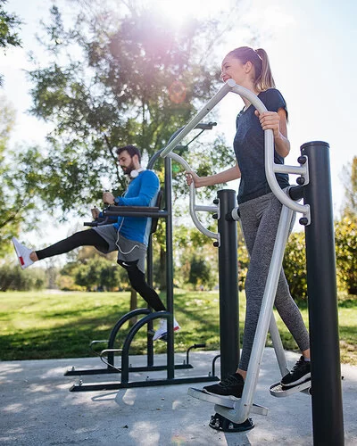 Ein junger Mann und eine junge Frau in Sportkleidung trainieren in einem Park an Fitnessgeräten.