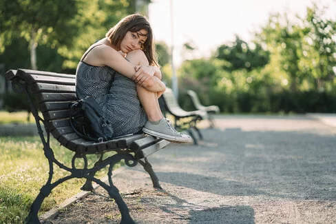 Eine junge Frau sitzt mit traurigem Gesichtsausdruck im Sommer auf einer Parkbank.