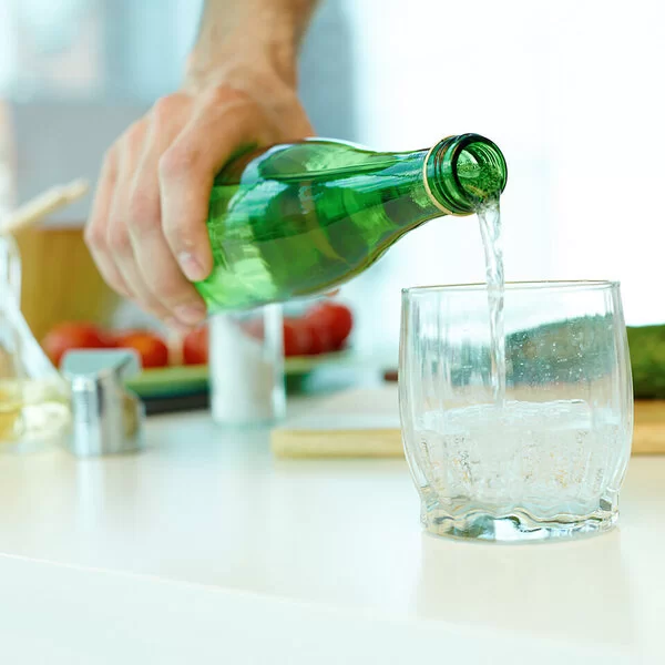 Ein Mann füllt Wasser aus einer grünen Glasflasche in ein Glas. Im Hintergrund zu sehen sind eine grüne Gurke, Olivenöl in einer Karaffe und Tomaten.