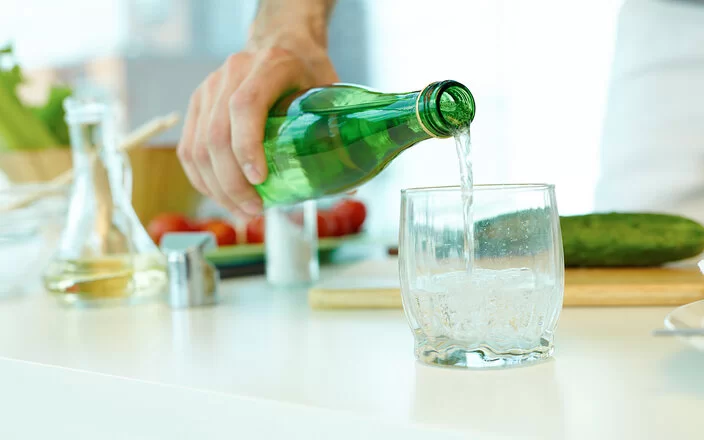 Ein Mann füllt Wasser aus einer grünen Glasflasche in ein Glas. Im Hintergrund zu sehen sind eine grüne Gurke, Olivenöl in einer Karaffe und Tomaten.