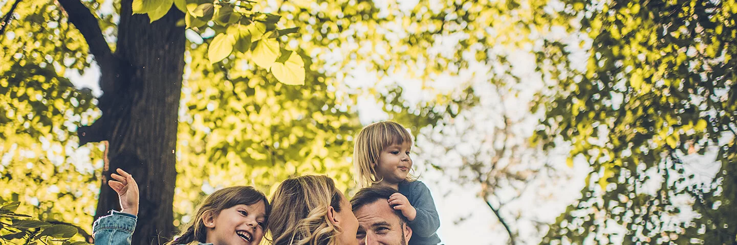 Eine junge vierköpfige Familie genießt unbeschwert einen Spaziergang im Wald. Die Eltern tragen die Kinder auf den Schultern.