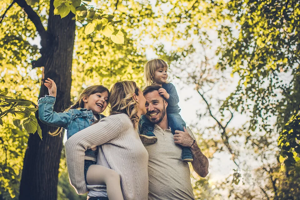 Eine junge vierköpfige Familie genießt unbeschwert einen Spaziergang im Wald. Die Eltern tragen die Kinder auf den Schultern.