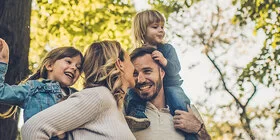 Eine junge vierköpfige Familie genießt unbeschwert einen Spaziergang im Wald. Die Eltern tragen die Kinder auf den Schultern.