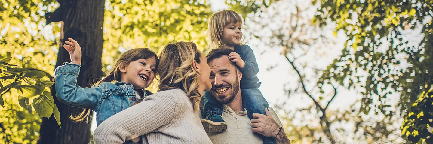 Eine junge vierköpfige Familie genießt unbeschwert einen Spaziergang im Wald. Die Eltern tragen die Kinder auf den Schultern.