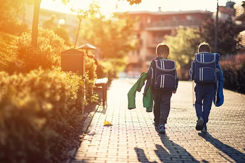 Zwei kleine Jungs gehen im hellen Sonnenlicht eine Spielstraße entlang, auf dem Rücken tragen sie große Schulranzen.